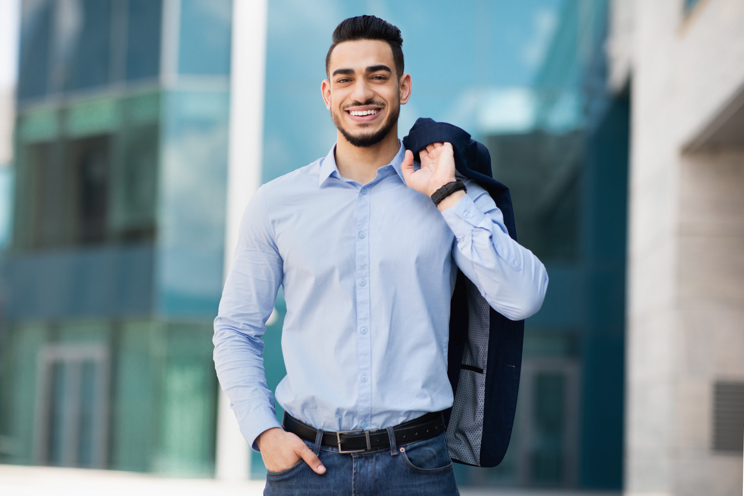 Confident businessman in a blue shirt holding a jacket over his shoulder, standing outside an office building.