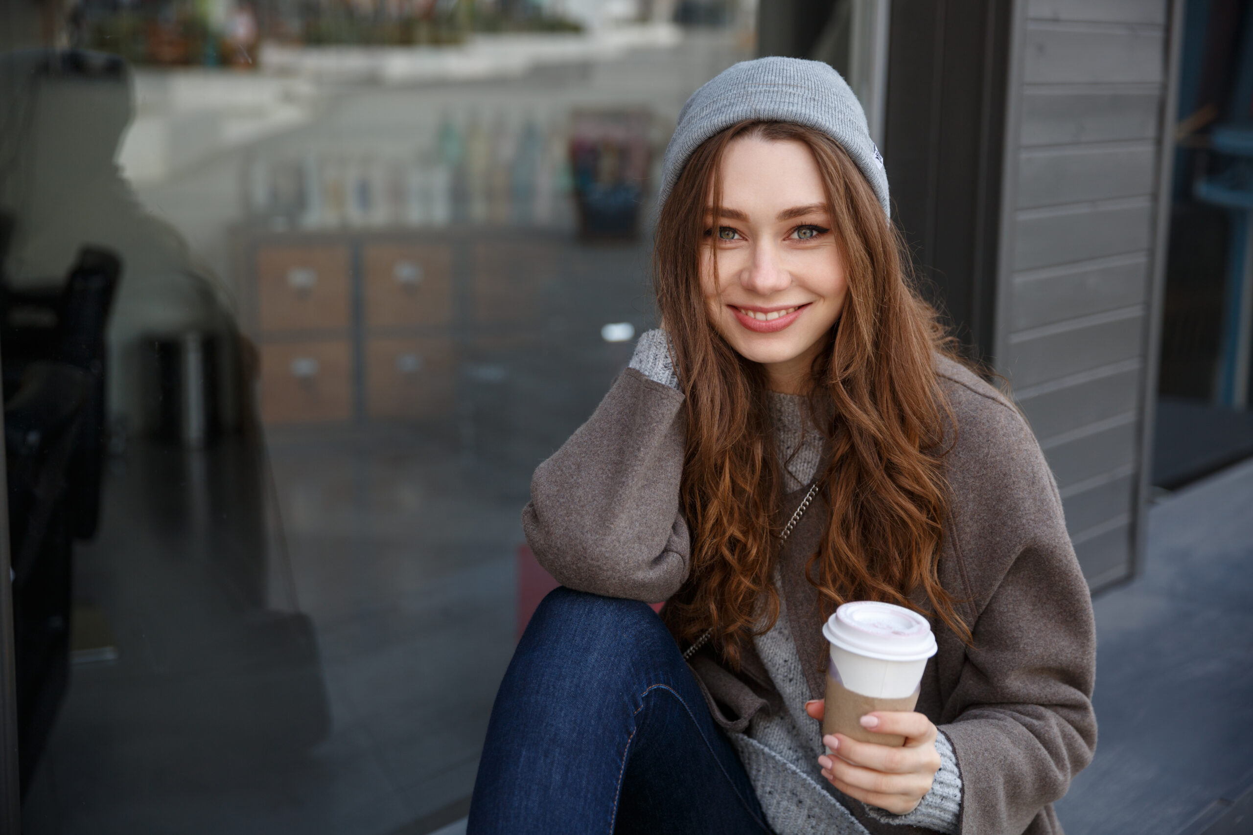 Smiling young woman drinking coffee to-go in the city, wearing a cozy sweater and beanie.